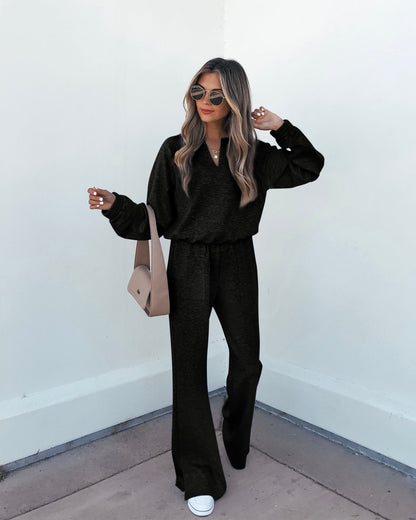 Woman in Stay Active Black Lounge Sweatpants, black top, white sneakers, sunglasses, and beige crossbody bag by a light wall.