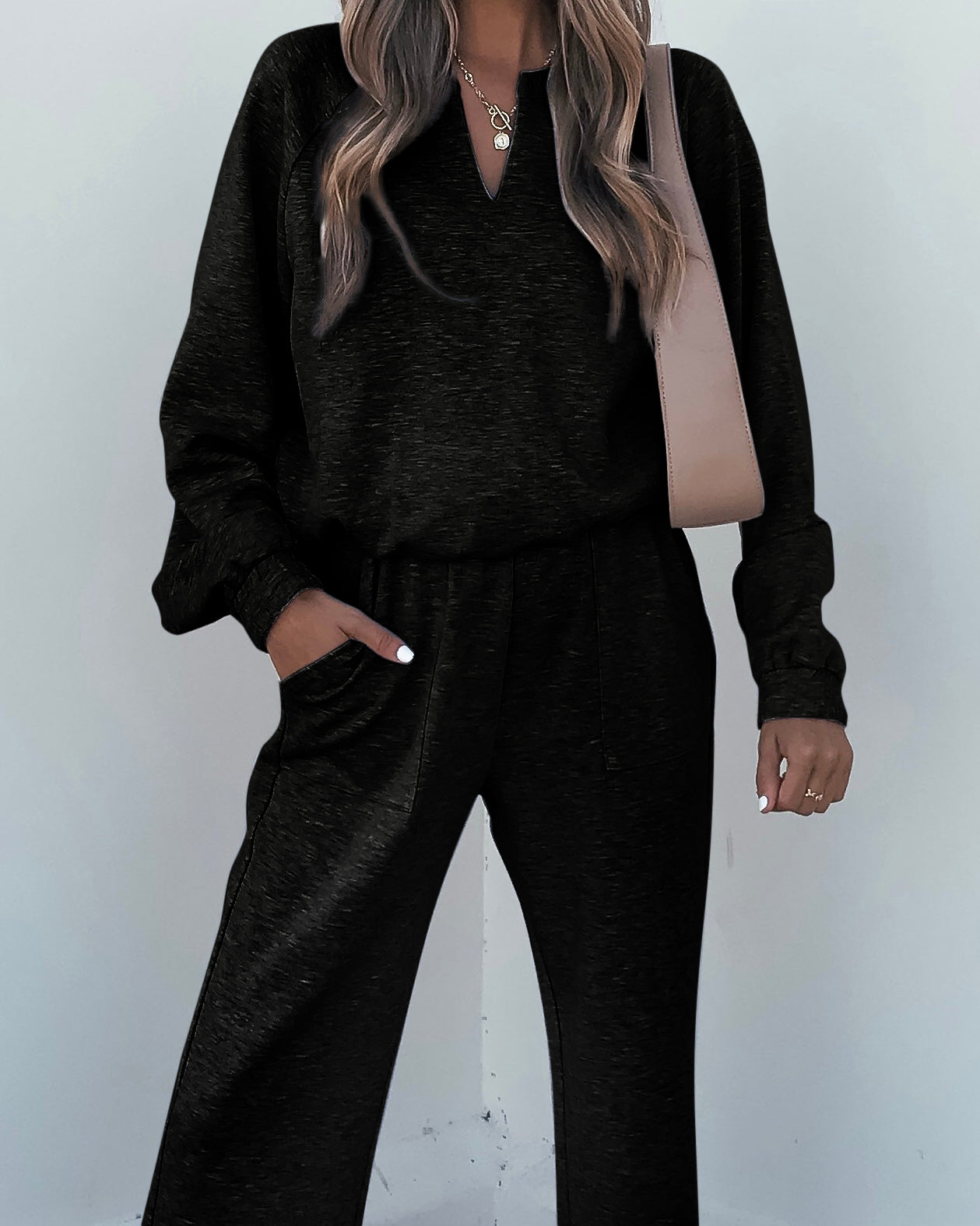 A woman in Stay Active Black Lounge Sweatpants, a loose black top, beige shoulder bag, and layered gold necklaces.