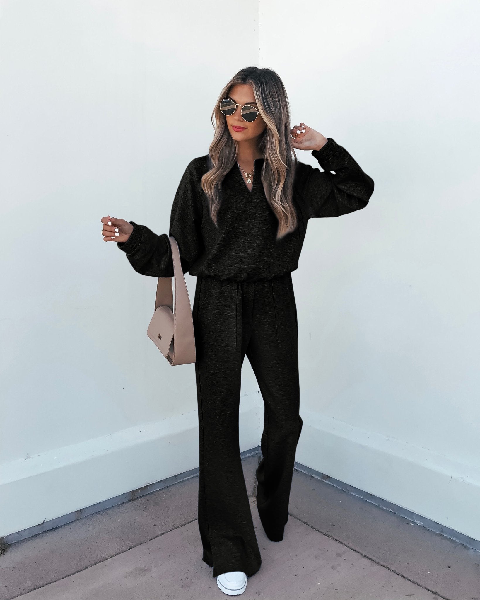 Woman in the Stay Active Black Lounge Sweatshirt, matching sweatpants, sneakers, sunglasses, and a crossbody bag stands by a white wall.