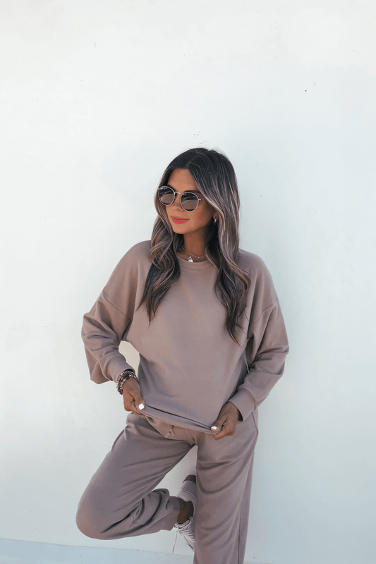 A woman standing against a white wall, wearing a mocha-colored Step Up Lounge Set consisting of a long-sleeve sweatshirt and matching joggers.