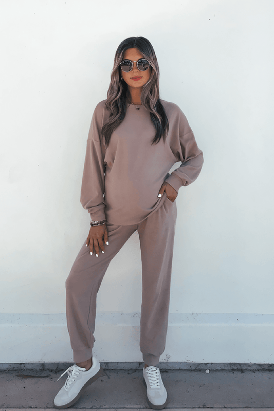 Woman in sunglasses wears the Step Up Lounge Set - Mocha with white sneakers, standing against a white wall, hand on hip.