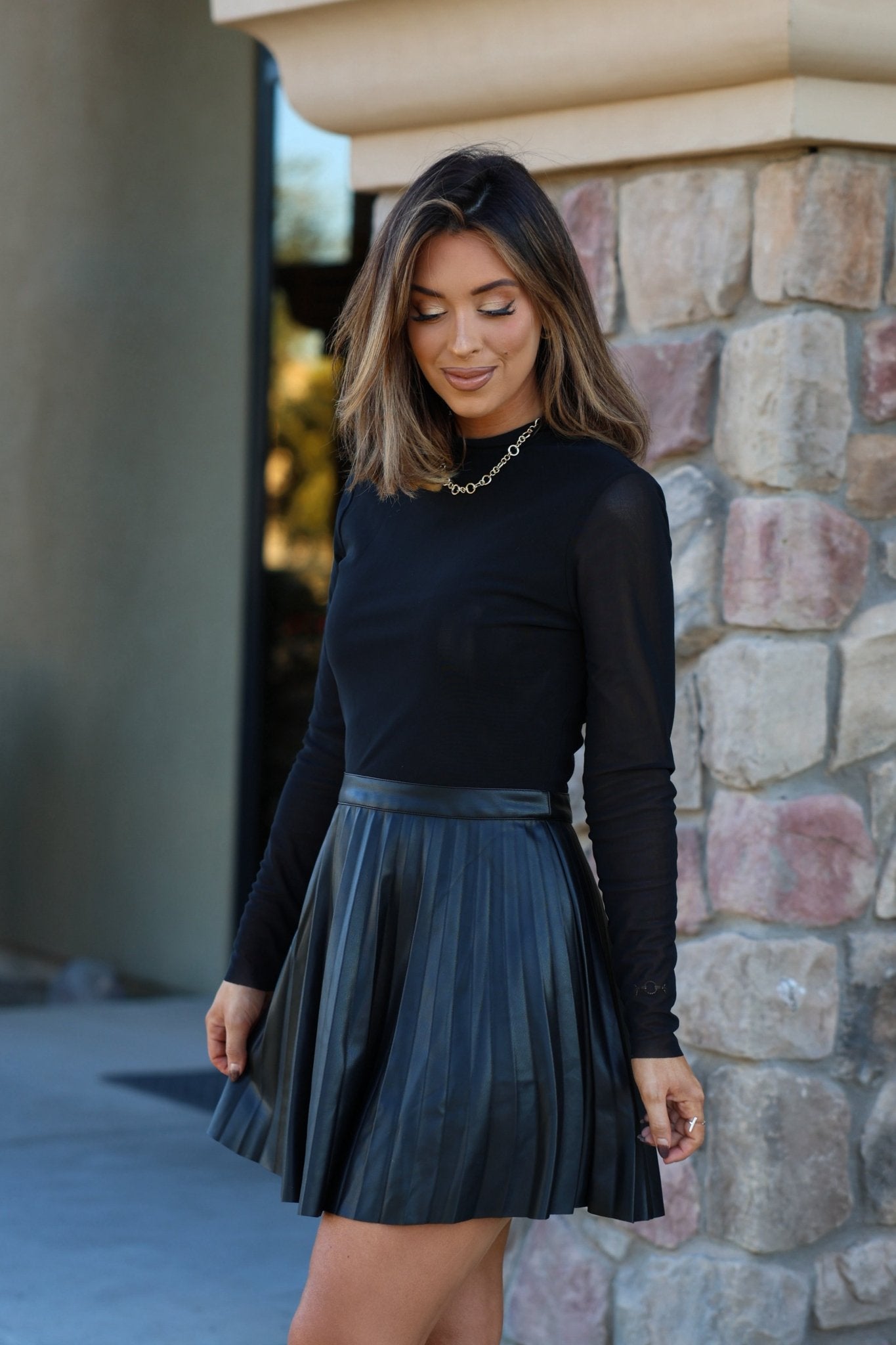 A woman in the Steve Madden Black Nevah Mini Dress stands by a stone wall, looking down and smiling.