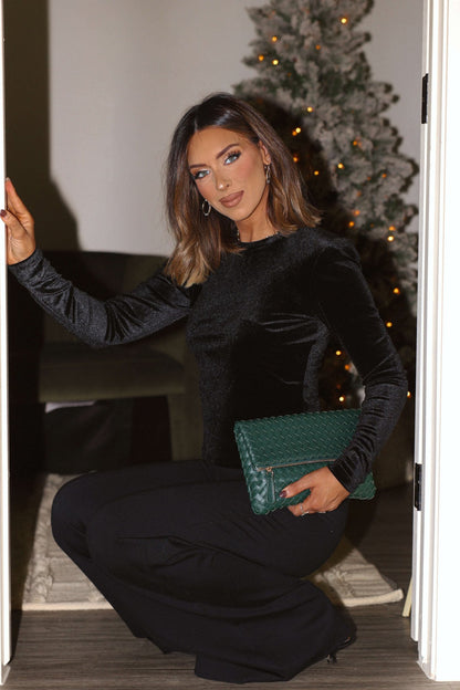 A woman wearing the Steve Madden Black Velvet Maddilynn Top kneels by an open door, holding a green clutch near a Christmas tree.