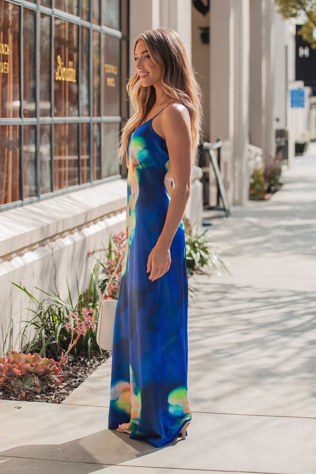 A woman in the Steve Madden Blue Atara Print Slip Maxi Dress stands on a sunny sidewalk with a white handbag and plants nearby.