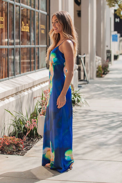 A woman in the Steve Madden Blue Atara Print Slip Maxi Dress stands on a sunny sidewalk with a white handbag and plants nearby.
