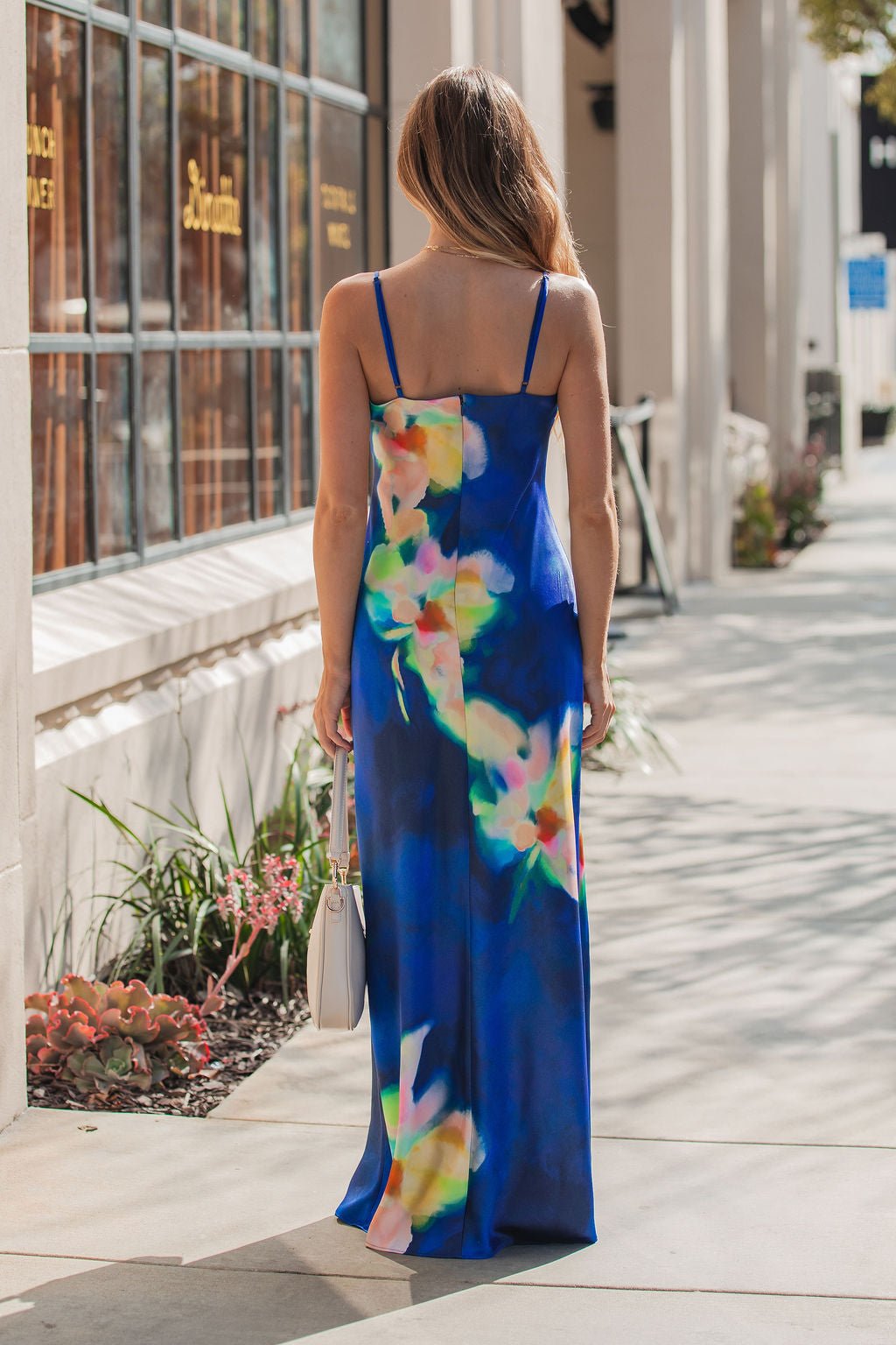 A woman in the Steve Madden Blue Atara Print Slip Maxi Dress stands on a sunny sidewalk, her back to the camera, holding a white handbag.