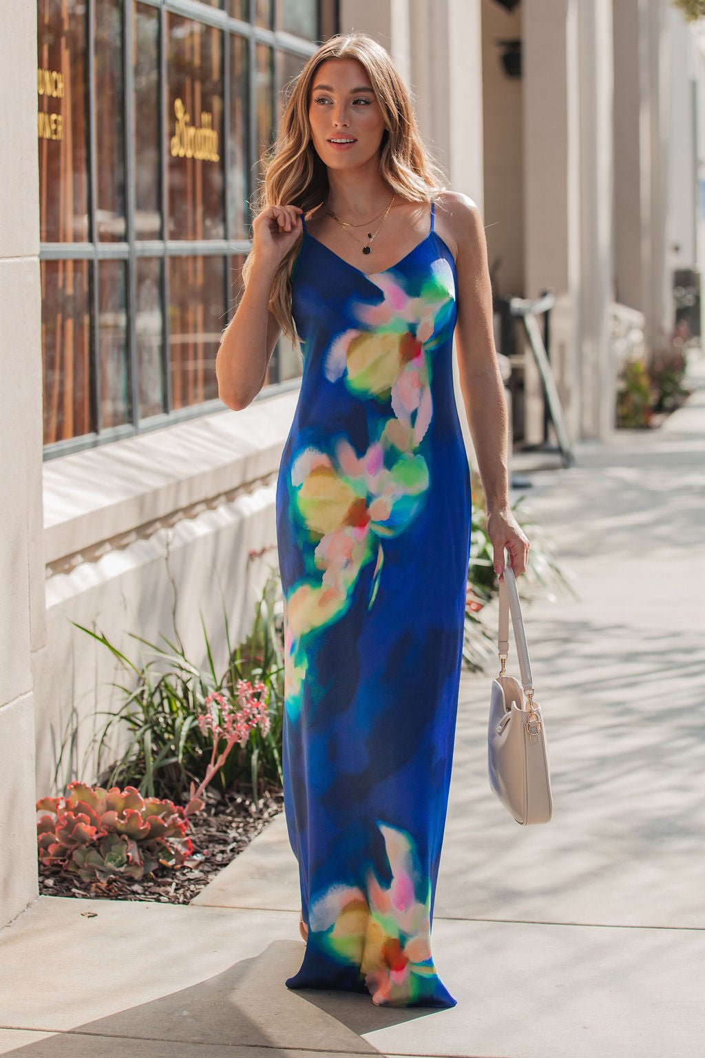 A woman walks on a city sidewalk in the Steve Madden Blue Atara Print Slip Maxi Dress, carrying a light handbag.