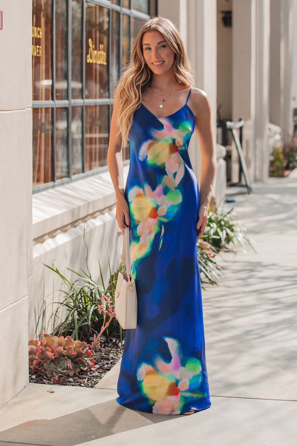 Woman in the Steve Madden Blue Atara Print Slip Maxi Dress stands outside by windows, smiling and holding a white purse.