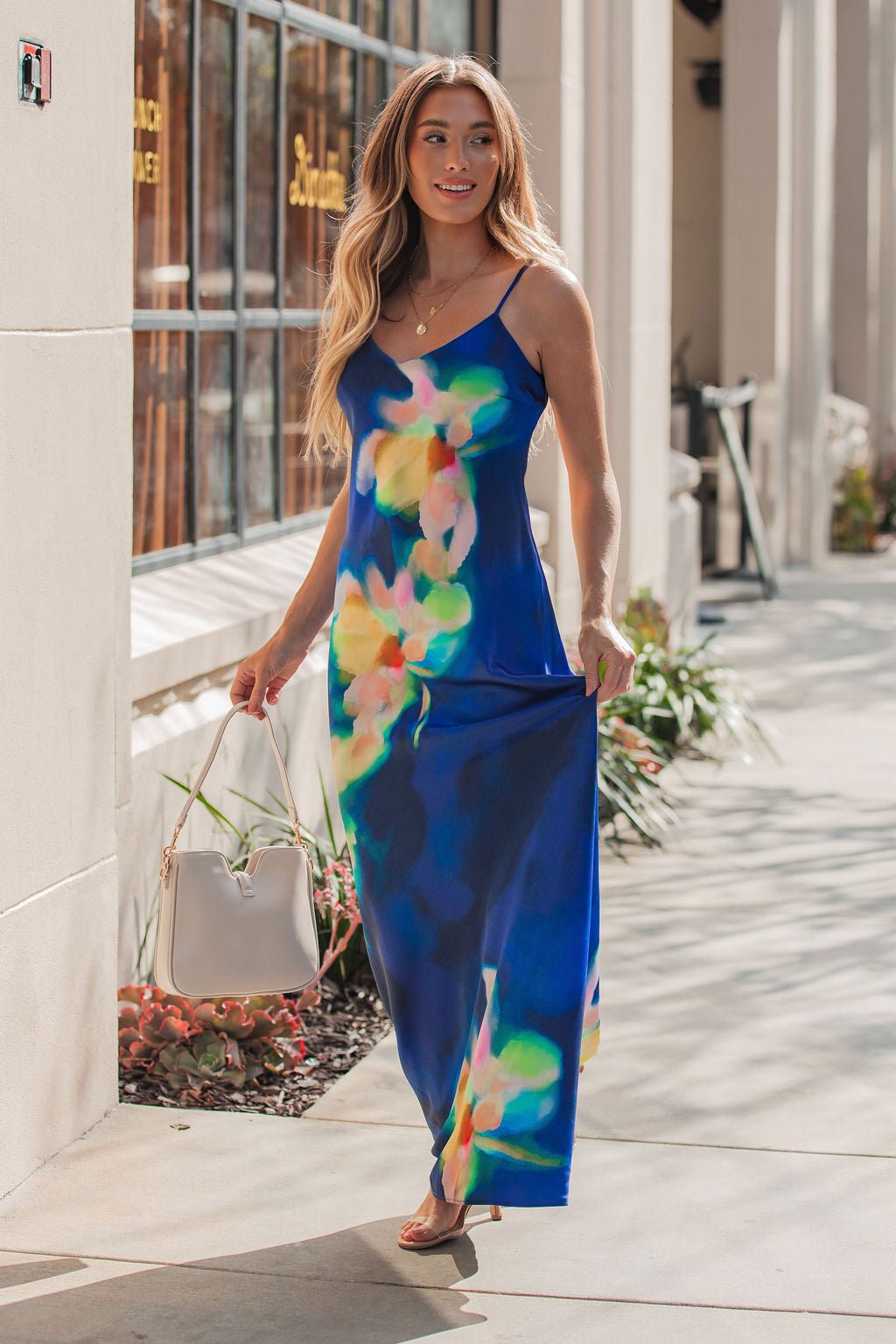 Woman in the Steve Madden Blue Atara Print Slip Maxi Dress walks on a sunlit sidewalk, holding a white handbag.