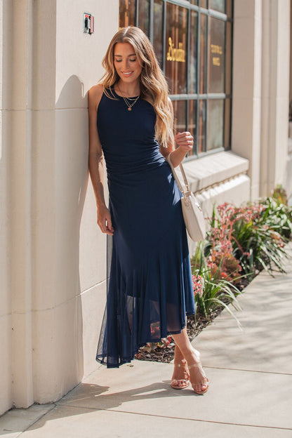 Smiling, she stands outside in a Steve Madden Navy Laney Asymmetrical Midi Dress, holding a white purse and wearing sandals.