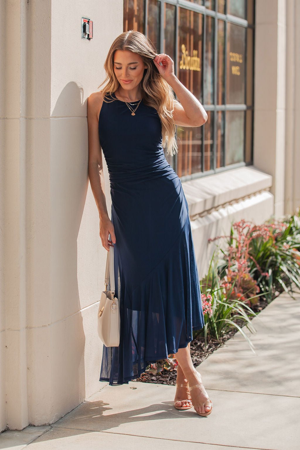 Woman wearing a Steve Madden Navy Laney Asymmetrical Midi Dress and sandals stands by a building, holding a white purse.