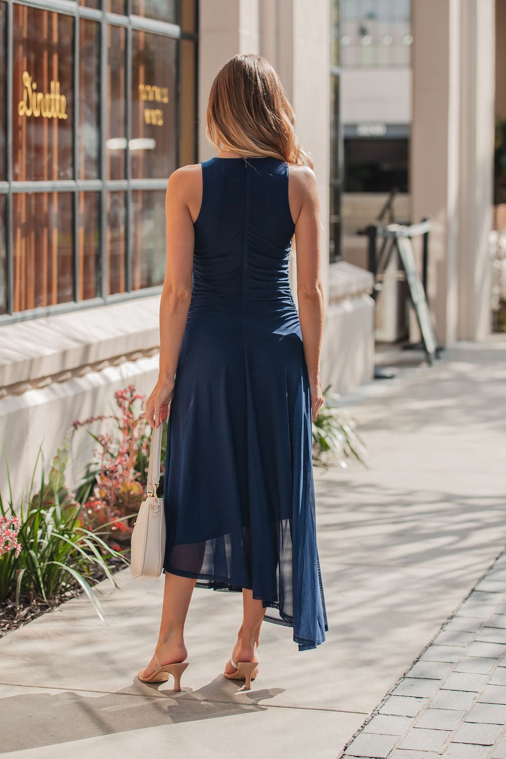 Woman in a sleeveless Steve Madden Navy Laney Asymmetrical Midi Dress and heels walks on the sidewalk, carrying a white handbag.