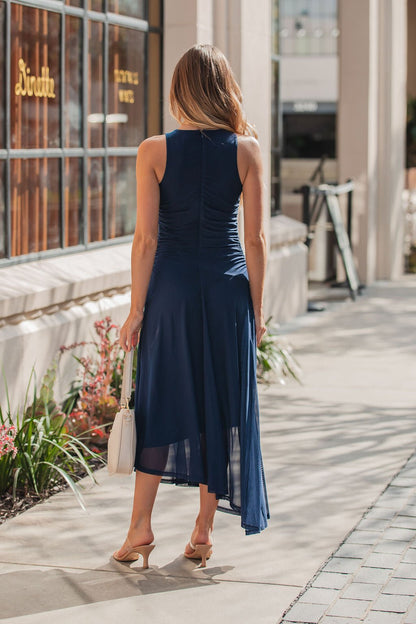 Woman in a sleeveless Steve Madden Navy Laney Asymmetrical Midi Dress and heels walks on the sidewalk, carrying a white handbag.