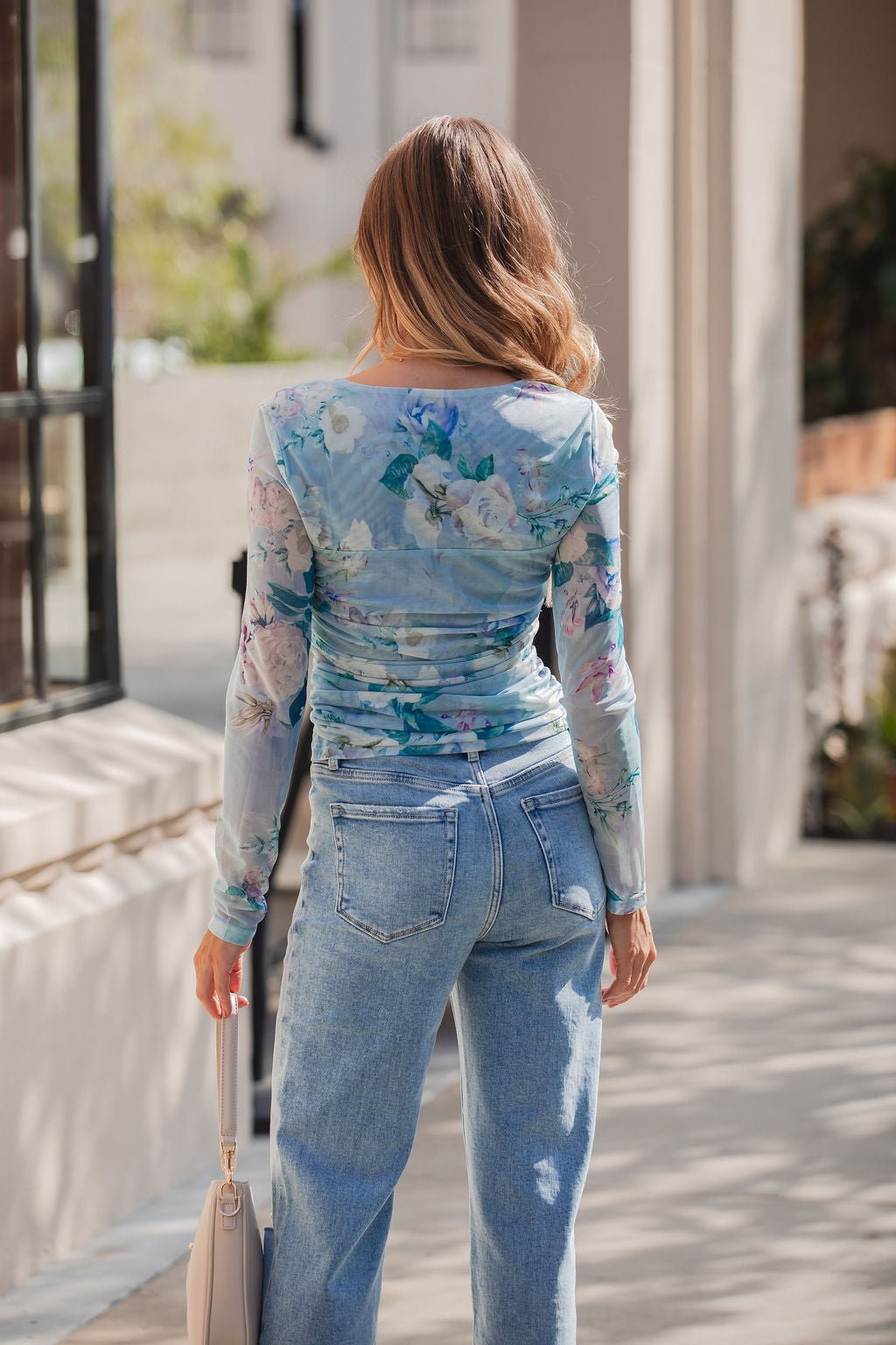 A woman with wavy hair wears the Steve Madden Blue Zinia Print Top and light blue jeans, standing outside facing away from the camera.