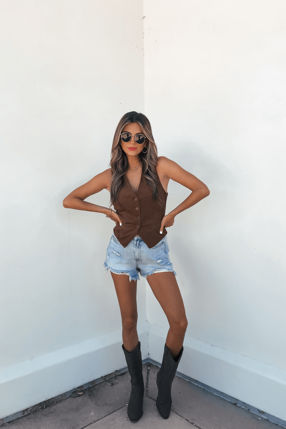 Woman in a Steve Madden Brown Barrett Vest - FINAL SALE, denim shorts, and boots stands confidently against a white wall, hands on hips.
