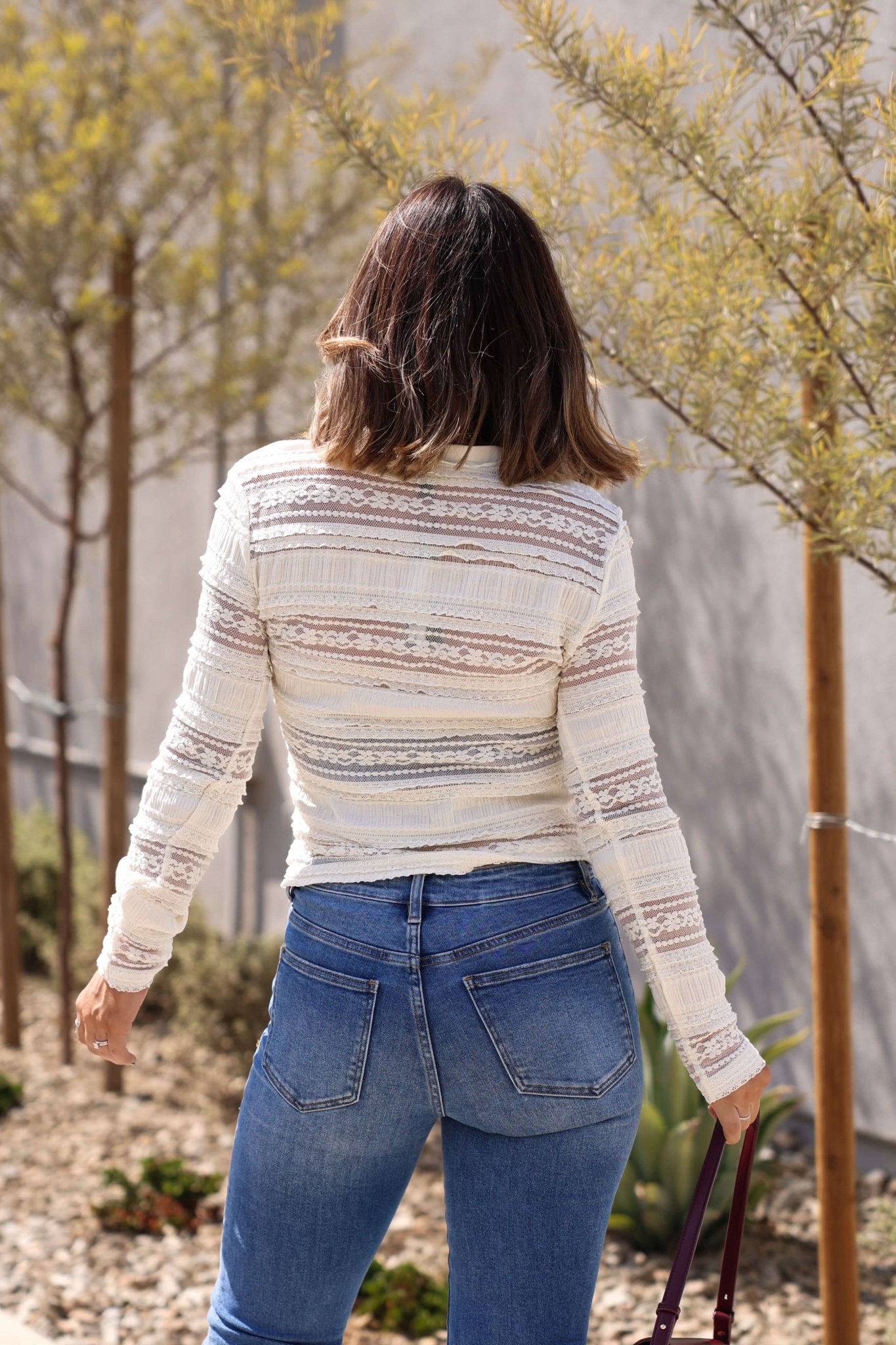 A woman in a Steve Madden Caterina Cream Lace Top and blue jeans walks outdoors near small trees and plants.