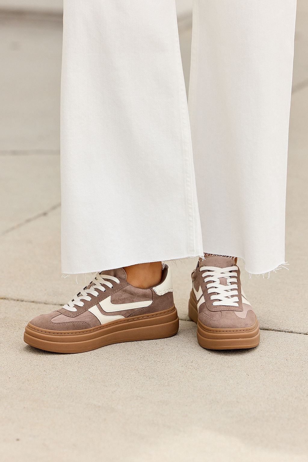Person in white wide-leg pants and taupe Steve Madden Dodge Sneakers with white laces stands on light pavement.