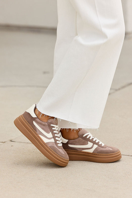 Person in white pants wears taupe Steve Madden Dodge Sneakers with round toe, walking on light concrete.