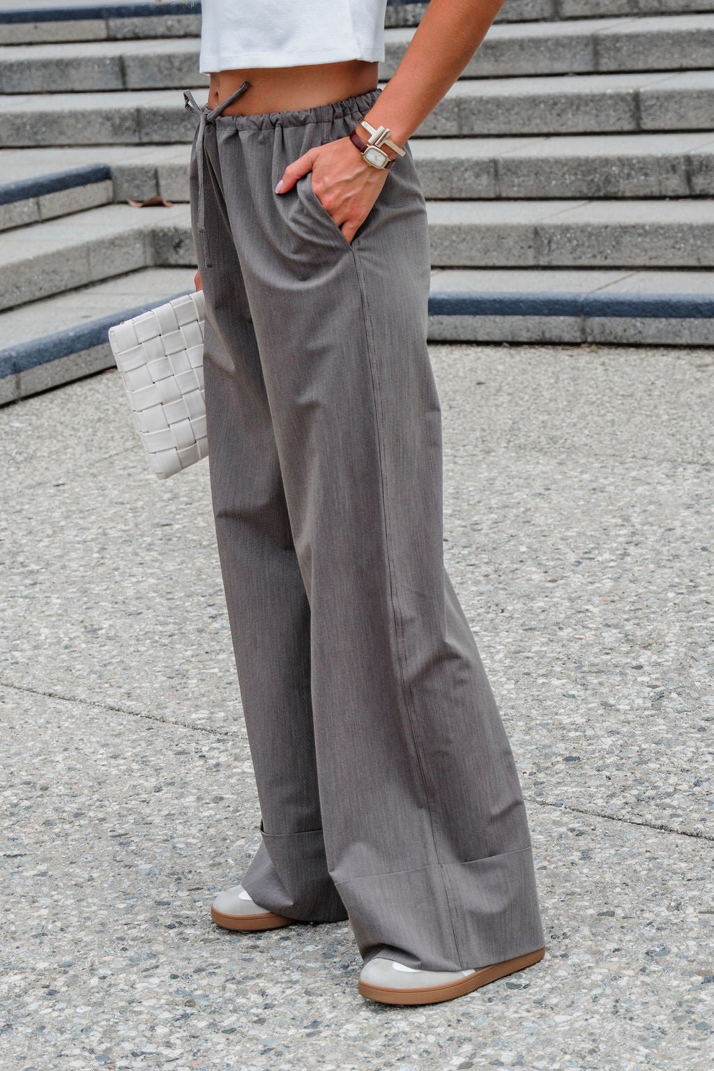Person standing on stone steps in Steve Madden Idra Pants - Brown, white top, tan shoes, watch, and holding a white woven clutch.