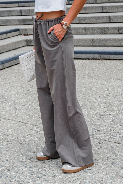Person standing on stone steps in Steve Madden Idra Pants - Brown, white top, tan shoes, watch, and holding a white woven clutch.