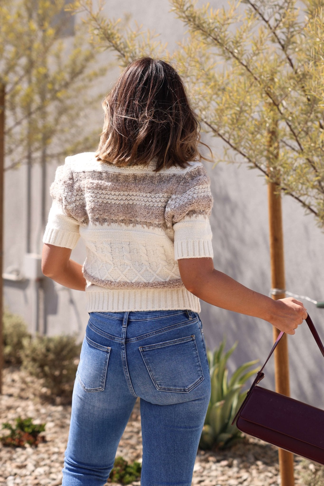 A woman with shoulder-length hair, in a Steve Madden Janae Cream Multi Sweater Cardigan and jeans, stands outdoors with a maroon bag.