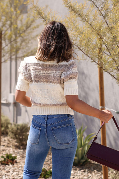 A woman with shoulder-length hair, in a Steve Madden Janae Cream Multi Sweater Cardigan and jeans, stands outdoors with a maroon bag.