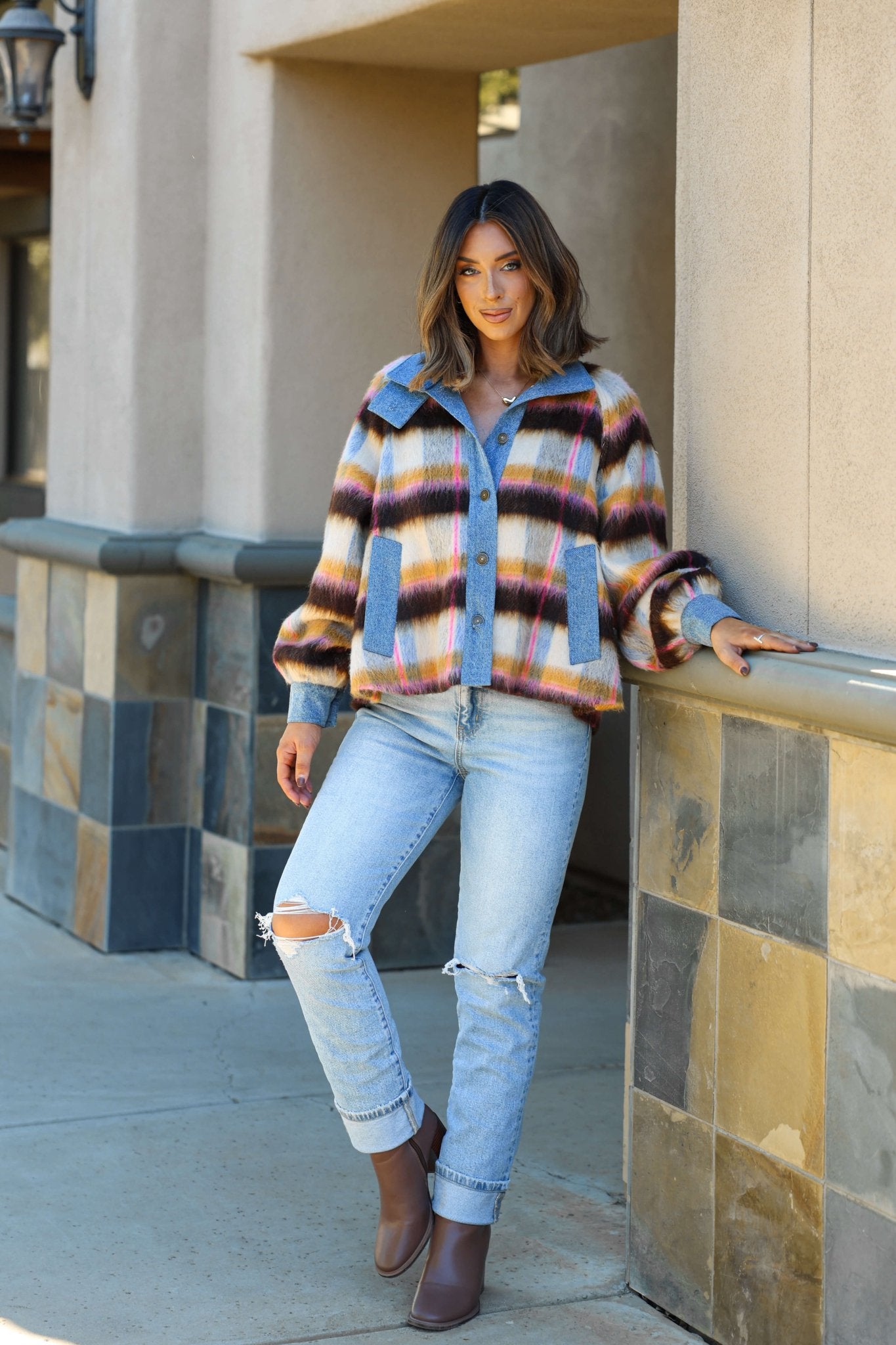 Woman in a relaxed fit Steve Madden Kaylen Multi Striped Jacket and ripped jeans smiles outdoors, leaning on a tiled wall.