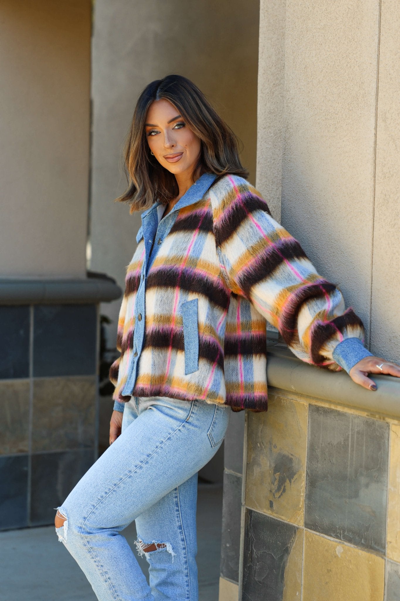 Woman in a Steve Madden Kaylen Multi Striped Jacket and ripped jeans leans casually against a stone wall, looking at the camera.
