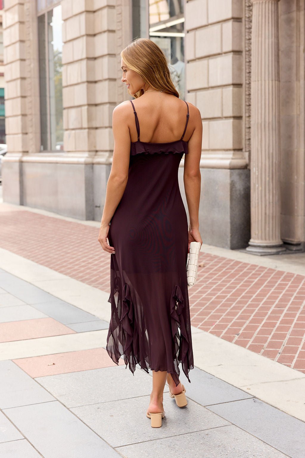Woman in a sleeveless brown Steve Madden Mariane Midi Dress with ruffled trim, beige heels, and a white clutch walks on a city sidewalk.