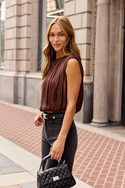 Woman with long wavy hair in a brown Steve Madden Milana Top, black jeans, holding a black purse on a city sidewalk.