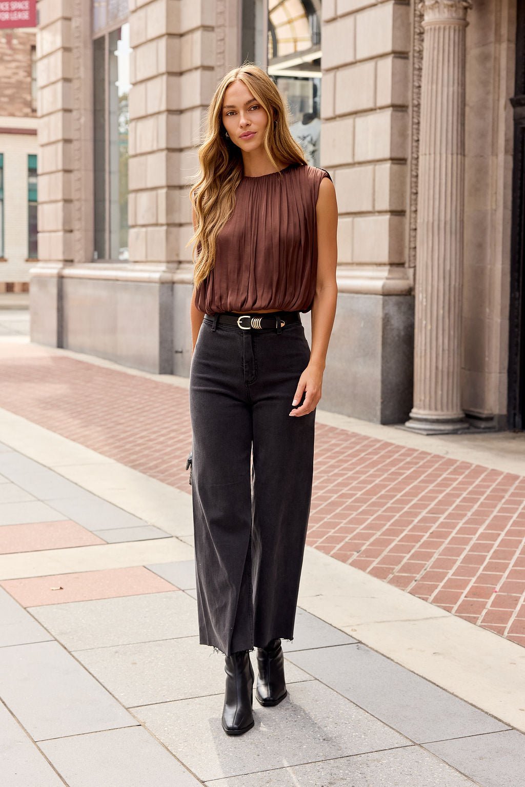 Woman with long hair in a brown Steve Madden Milana Top, black wide-leg pants, and boots walking by stone buildings.