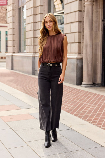 Woman with long hair in a brown Steve Madden Milana Top, black wide-leg pants, and boots walking by stone buildings.
