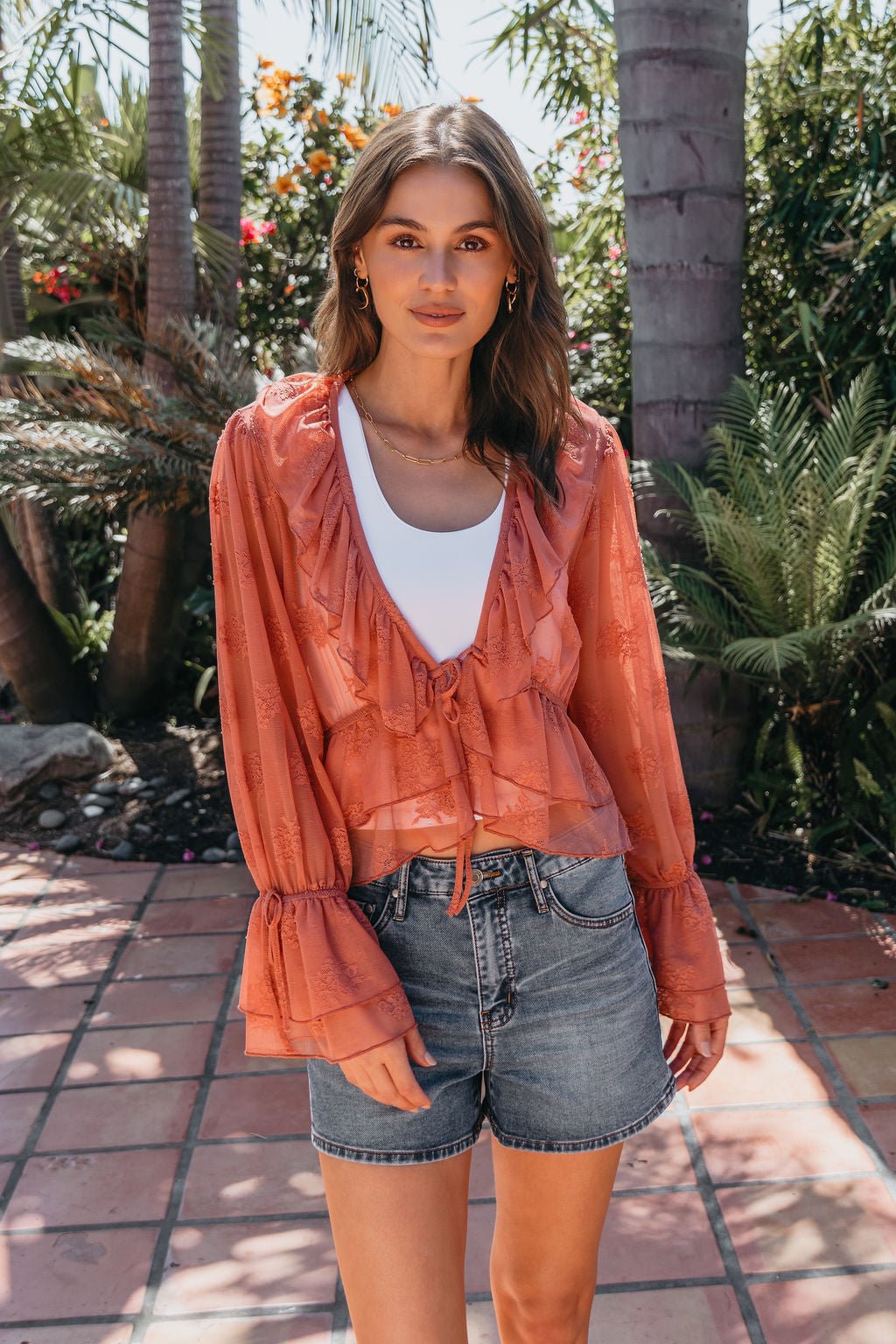 A woman stands outdoors in sunlight, wearing a Steve Madden Montana Rose Floral Top over a white top and denim shorts.