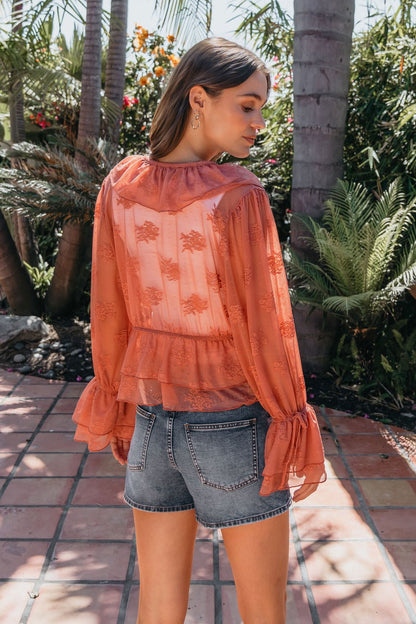 Woman stands outdoors in a garden, wearing the sheer Steve Madden Montana Rose Floral Top with ruffles and denim shorts, facing away.