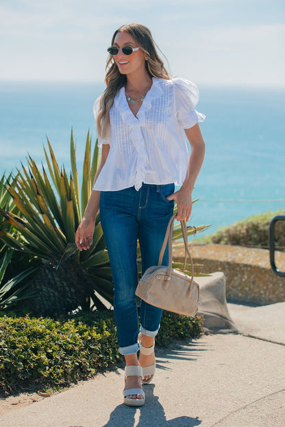 Woman in sunglasses, jeans, and a Steve Madden Natural Lyric Top holds a beige bag while walking along a sunny seaside path.