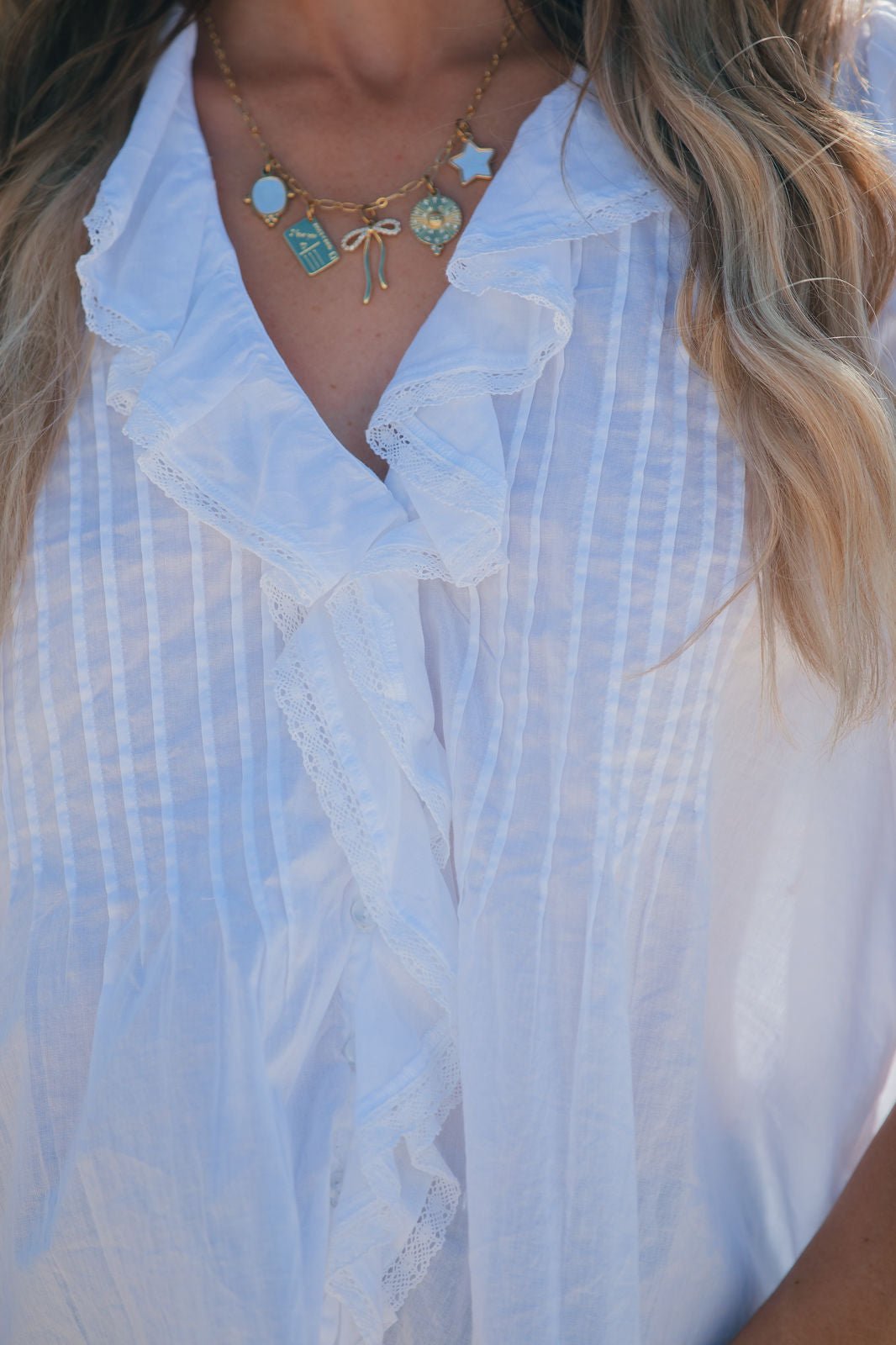 Wearing a ruffled Steve Madden Natural Lyric Top and a gold necklace with colorful charms, shown neck to mid-torso.