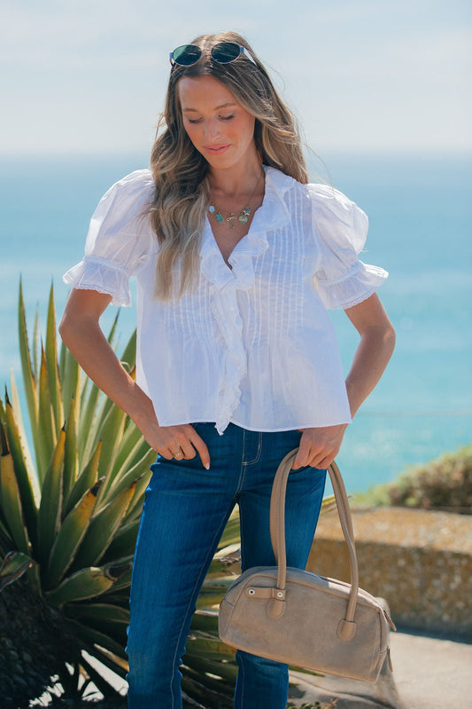 Woman in a Steve Madden Natural Lyric Top and jeans stands outdoors by the ocean, holding a beige handbag with plants behind her.