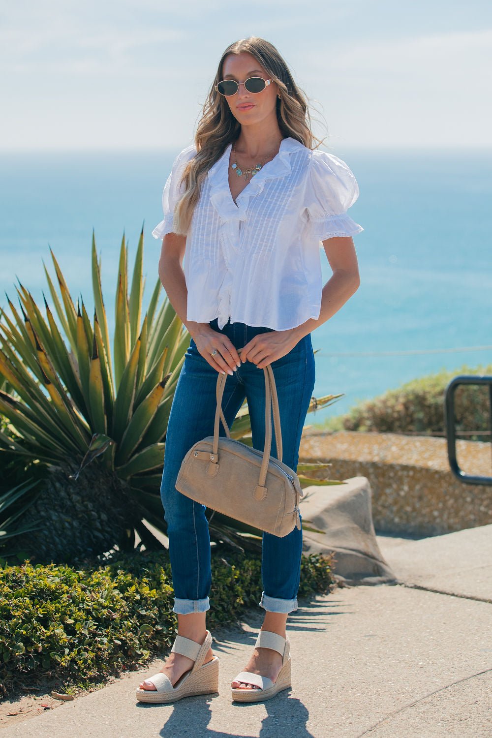 Woman in sunglasses, Steve Madden Natural Lyric Top, and jeans stands by the ocean with a tan handbag and wedge sandals.
