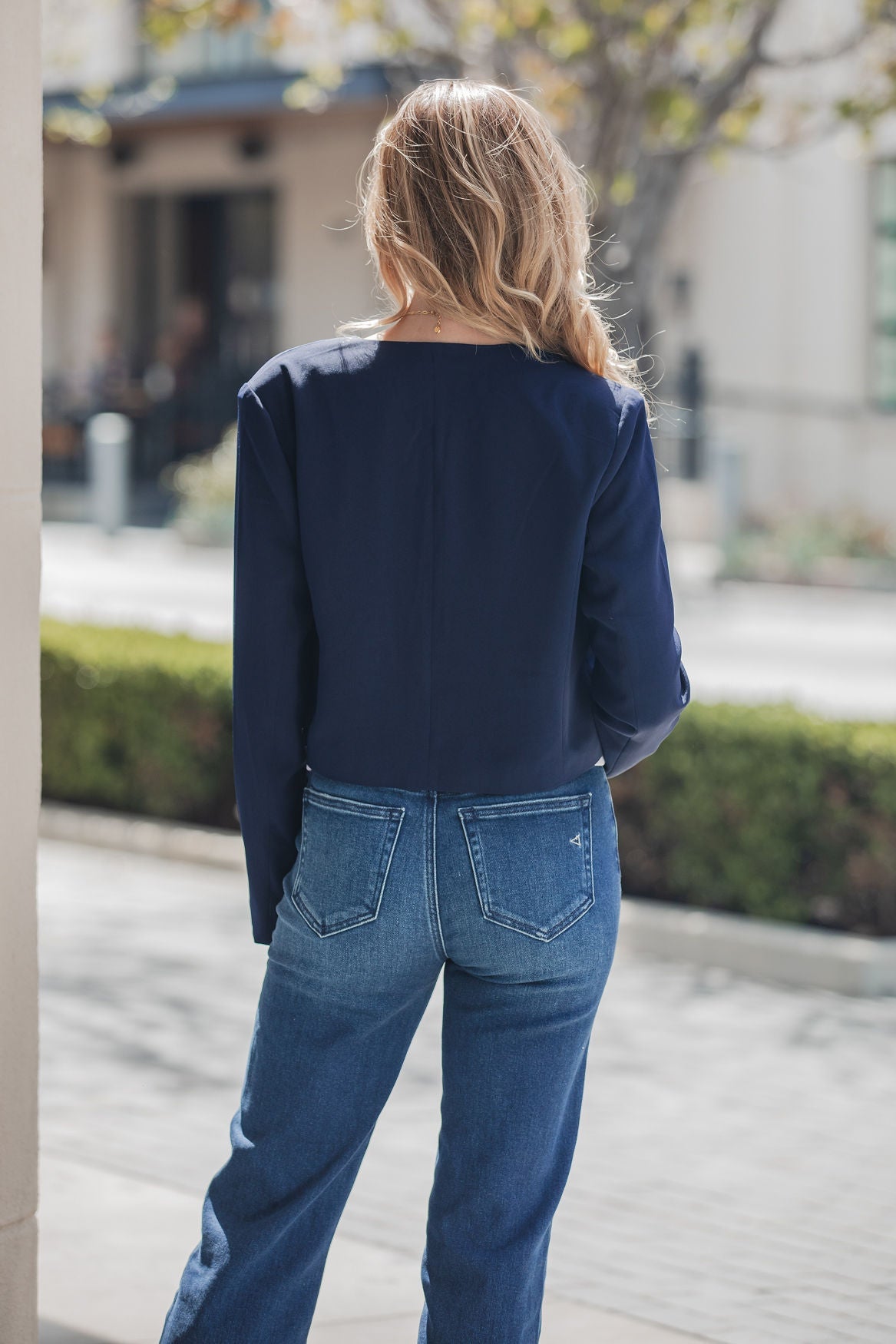 A woman with long blonde hair wears the Steve Madden Serena Blazer in navy, standing outdoors with her back to the camera.