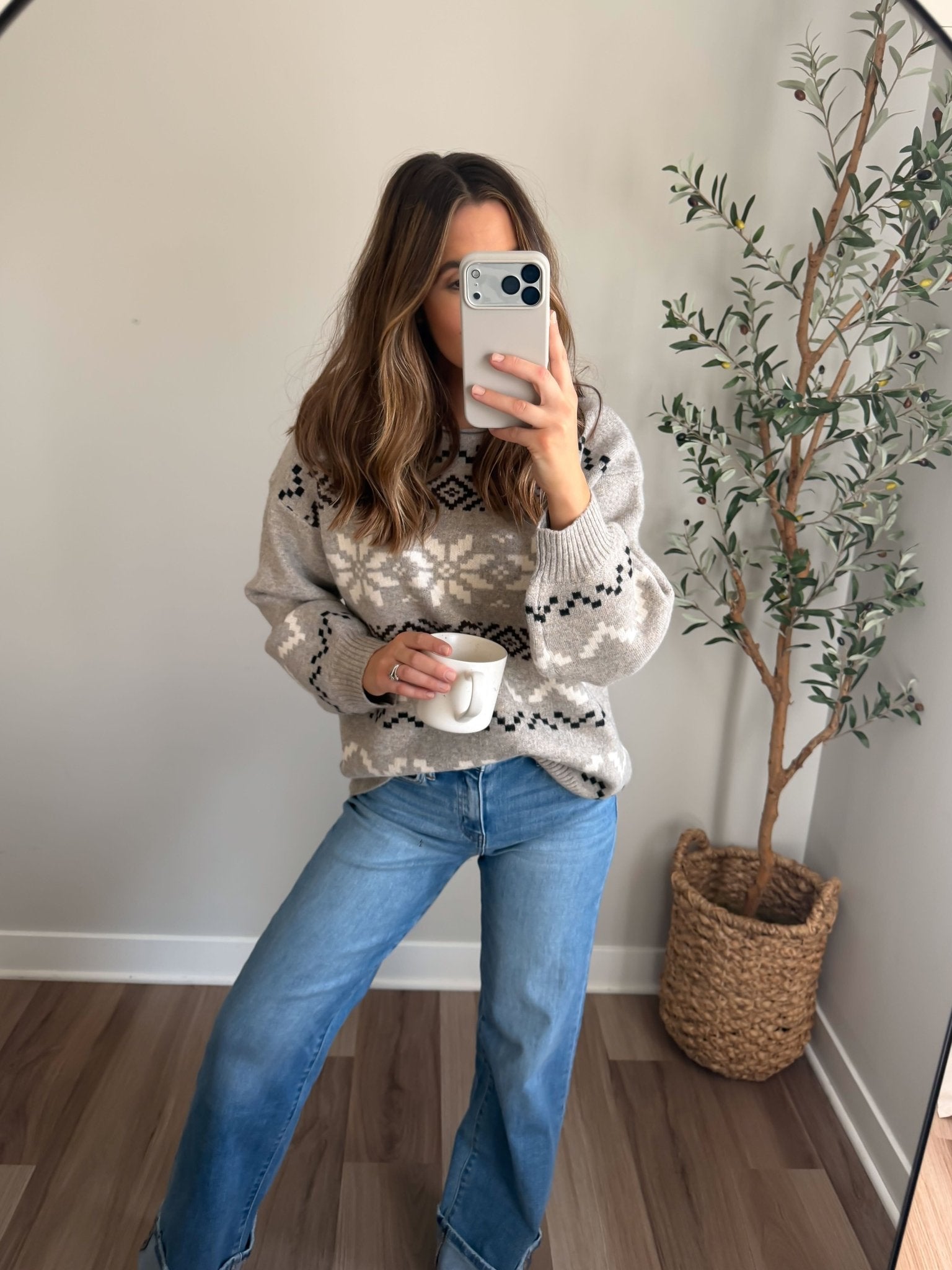 Woman taking a mirror selfie in a Stone Grey Snowflake Sweater and jeans, holding a white mug with a potted plant behind her.