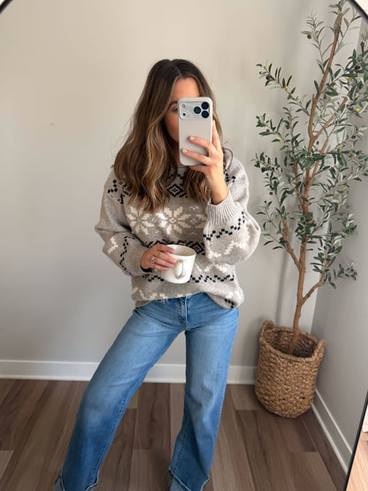 Woman taking a mirror selfie in a Stone Grey Snowflake Sweater and jeans, holding a white mug with a potted plant behind her.