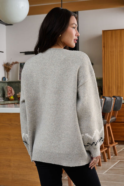 A woman with long dark hair wears a Stone Grey Snowflake Sweater in a modern kitchen, its winter pattern visible from behind.