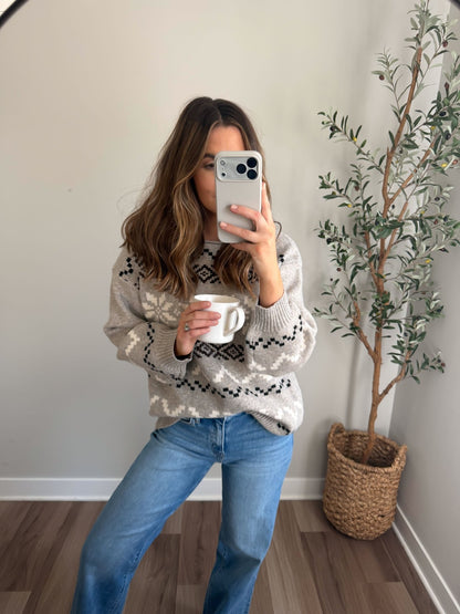 A woman in the Stone Grey Snowflake Sweater and jeans takes a mirror selfie with a white mug near a potted plant.