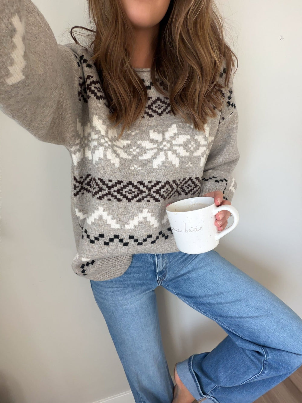 A woman in the Stone Grey Snowflake Sweater and jeans holds a white mug, standing barefoot against a neutral wall.