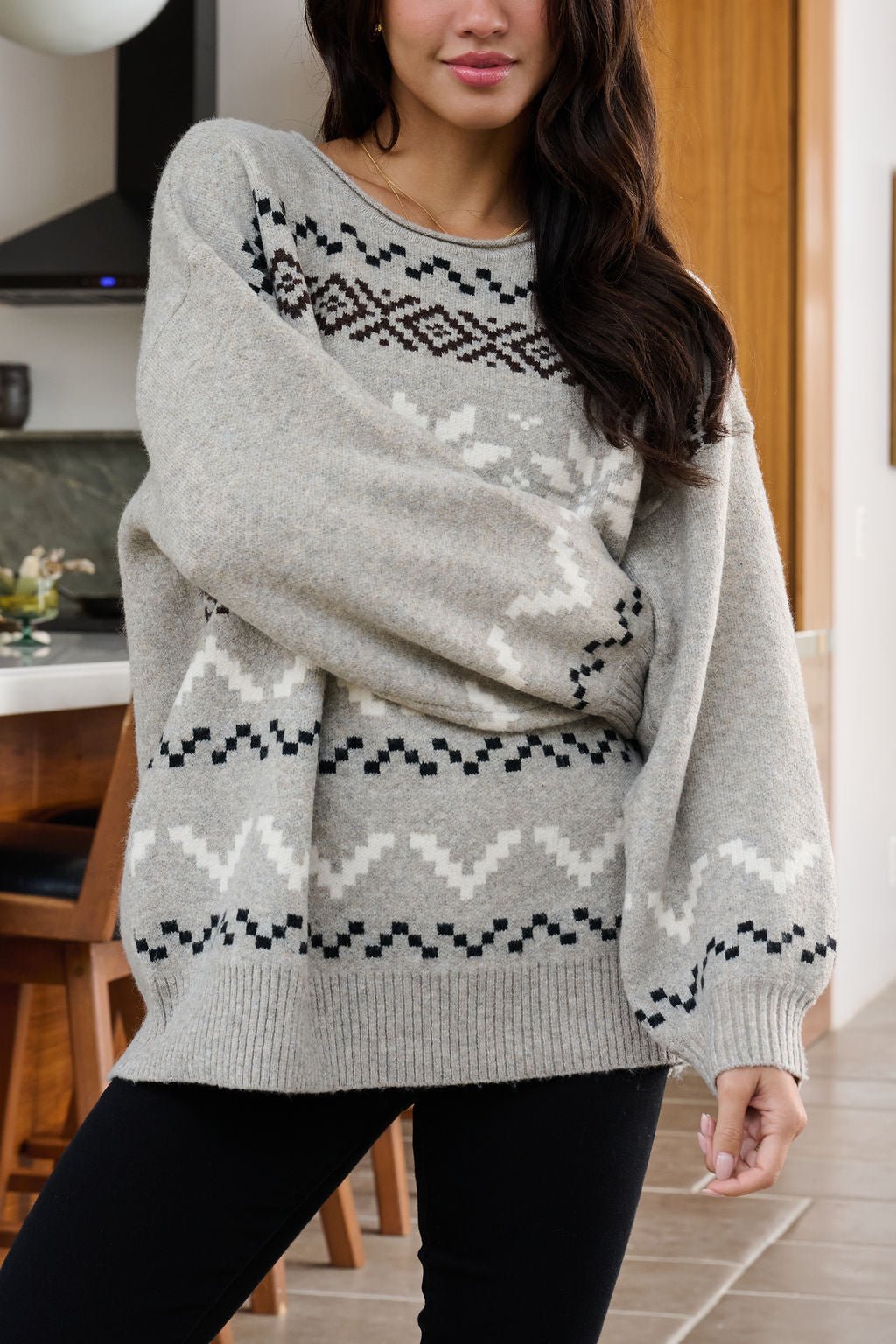 Woman in an oversized Stone Grey Snowflake Sweater stands indoors with wooden furniture in the background.