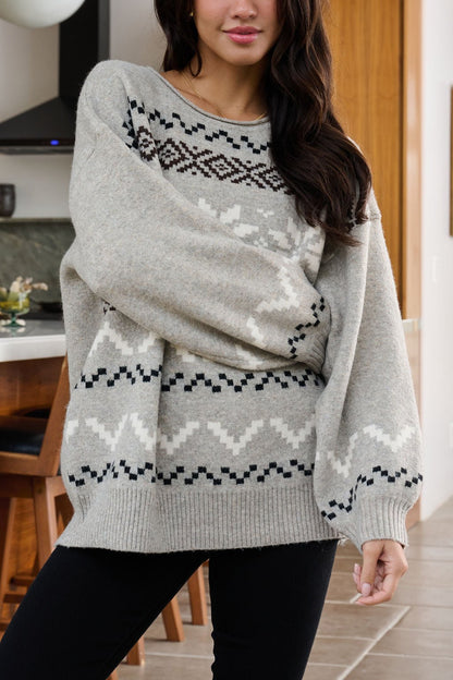 Woman in an oversized Stone Grey Snowflake Sweater stands indoors with wooden furniture in the background.