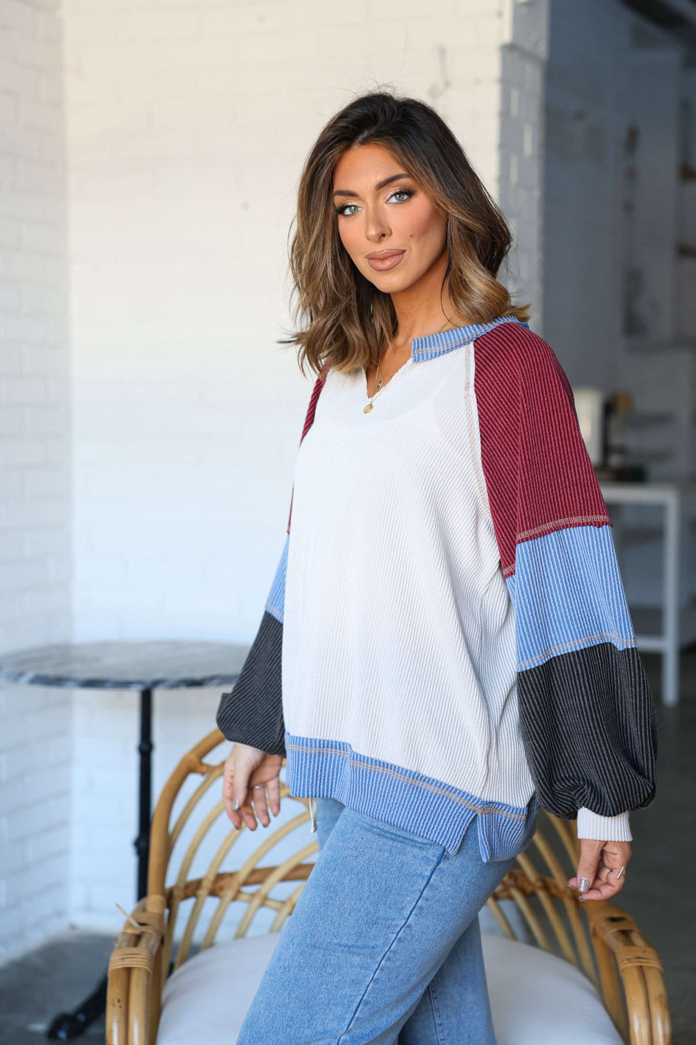Woman wears the Stone Multi Color Block Ribbed Pullover-FINAL SALE and blue jeans, sitting on a wicker chair in a bright room.