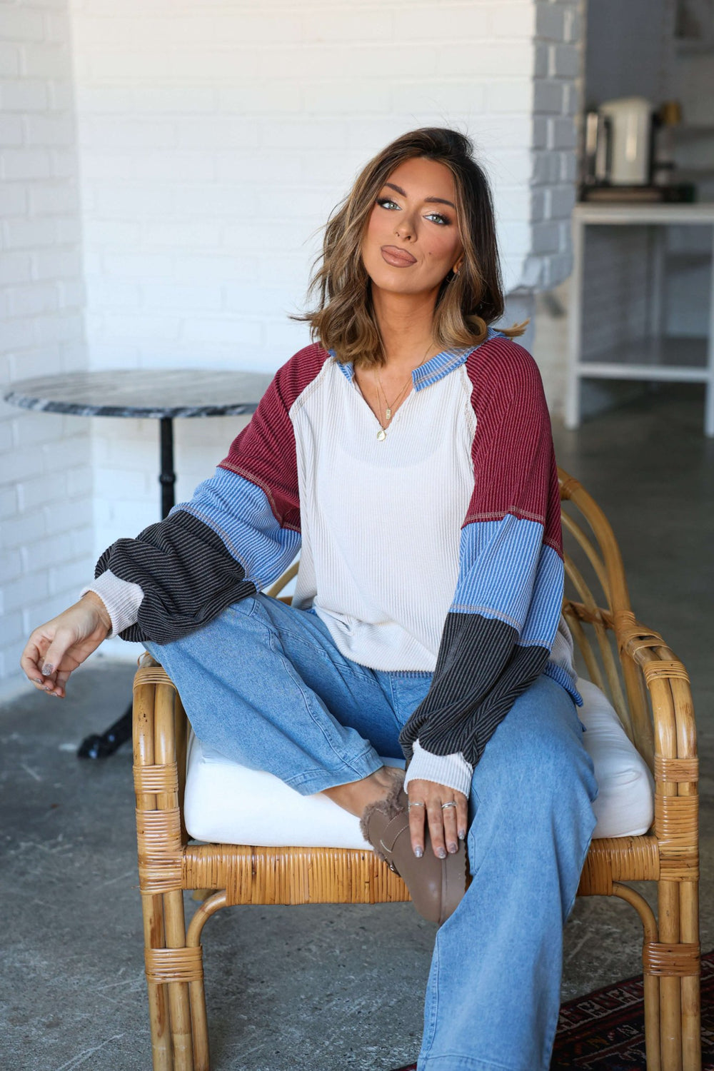 Woman with shoulder-length hair sits in a wicker chair, wearing the Stone Multi Color Block Ribbed Pullover and blue jeans.