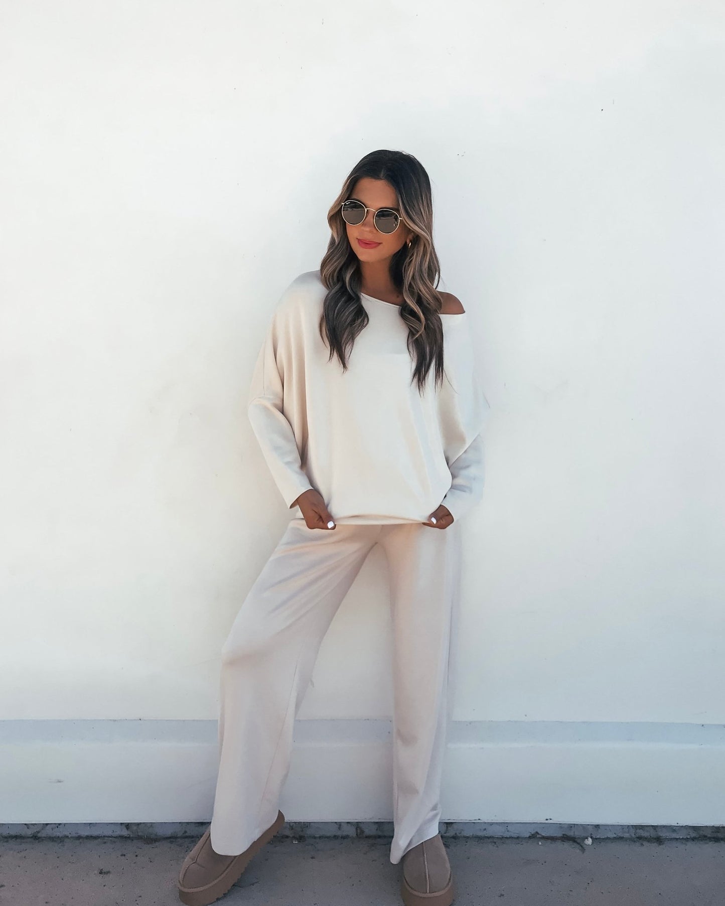 Woman in a Stone Off The Shoulder Scuba Top and sunglasses poses casually against a white wall with hands in pockets.