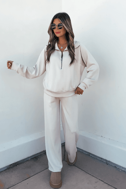 Woman wearing sunglasses, a light beige zip-up, Stone Soft Cloud Scuba Sweatpants, and beige shoes against a white wall.